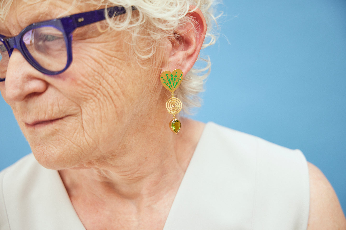 image of large firework heart earrings in green and gold on model with blue glasses and white hair against light blue background