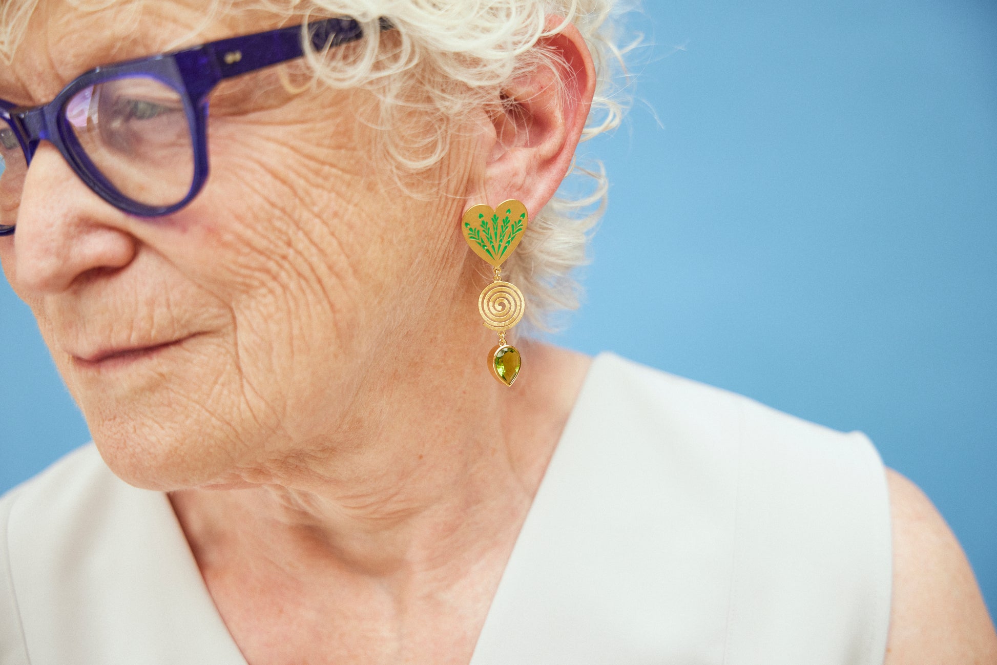 image of large firework heart earrings in green and gold on model with blue glasses and white hair against light blue background