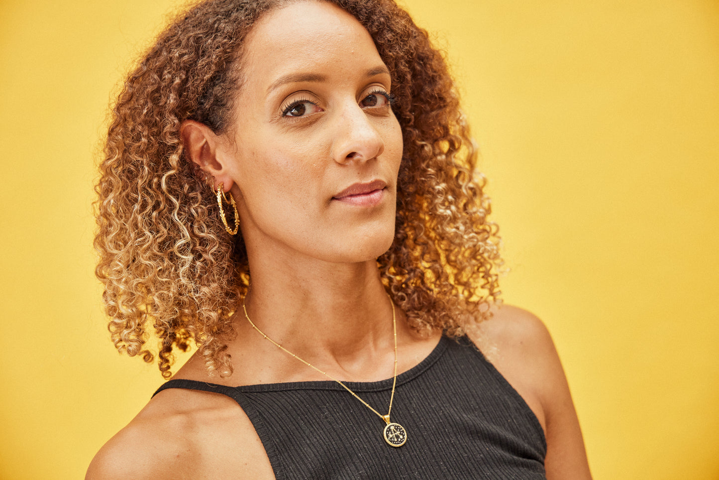 image of diamond sparkler initial necklace, letter A on short chain, on model with curly brown hair and brown skin against yellow background