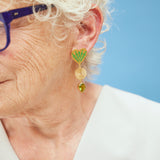 image of large firework heart earrings in green and gold on model with blue glasses and white hair against light blue background