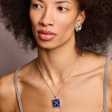 A model wearing a necklace with a square pendant featuring a silver emblem on a lapis lazuli background, surrounded by blue sapphires, and a chain attached to the top.