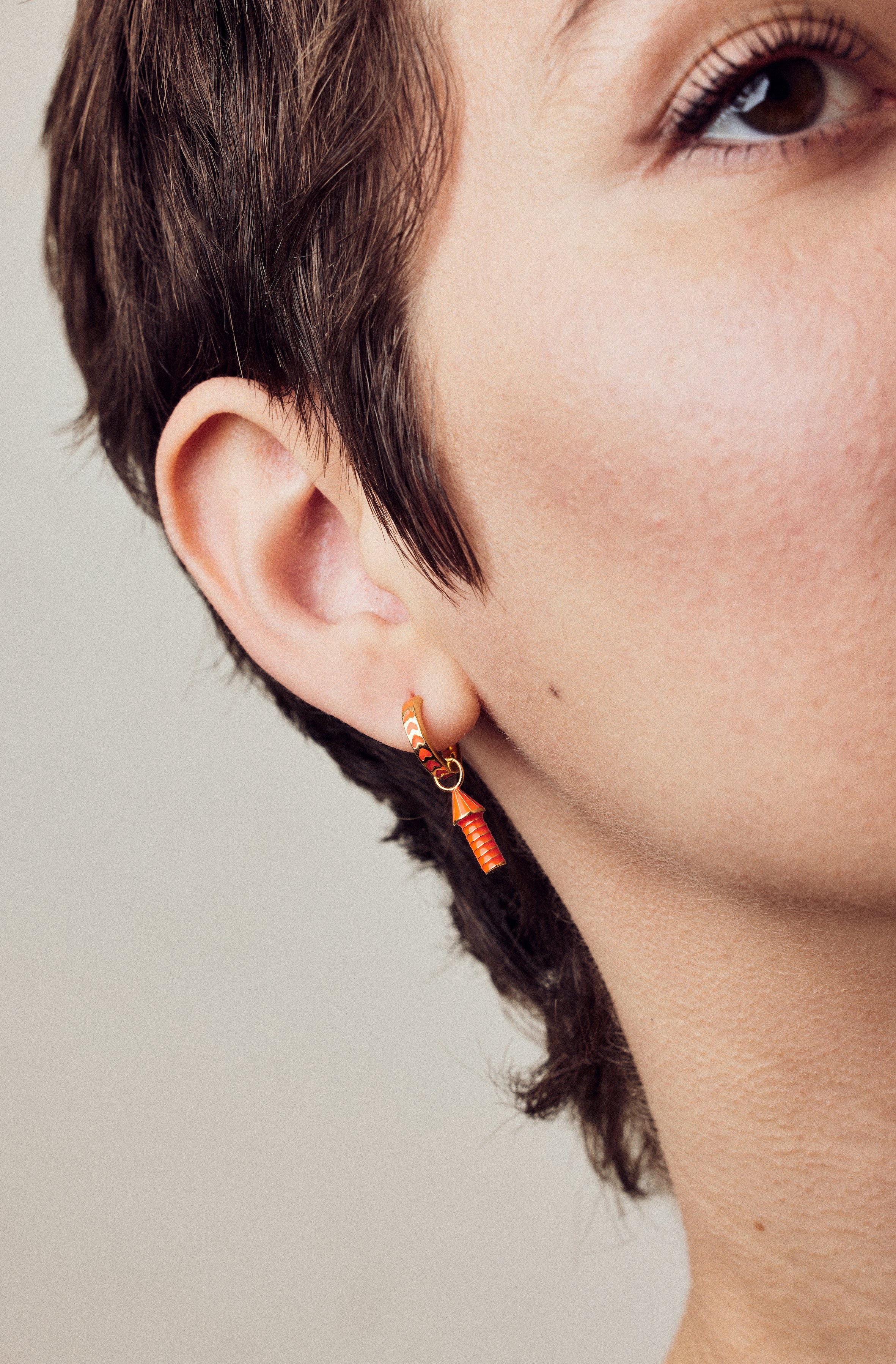 image of rocket enamel earrings in orange and gold shown close up of ear and eye on model with white skin and short brown hair
