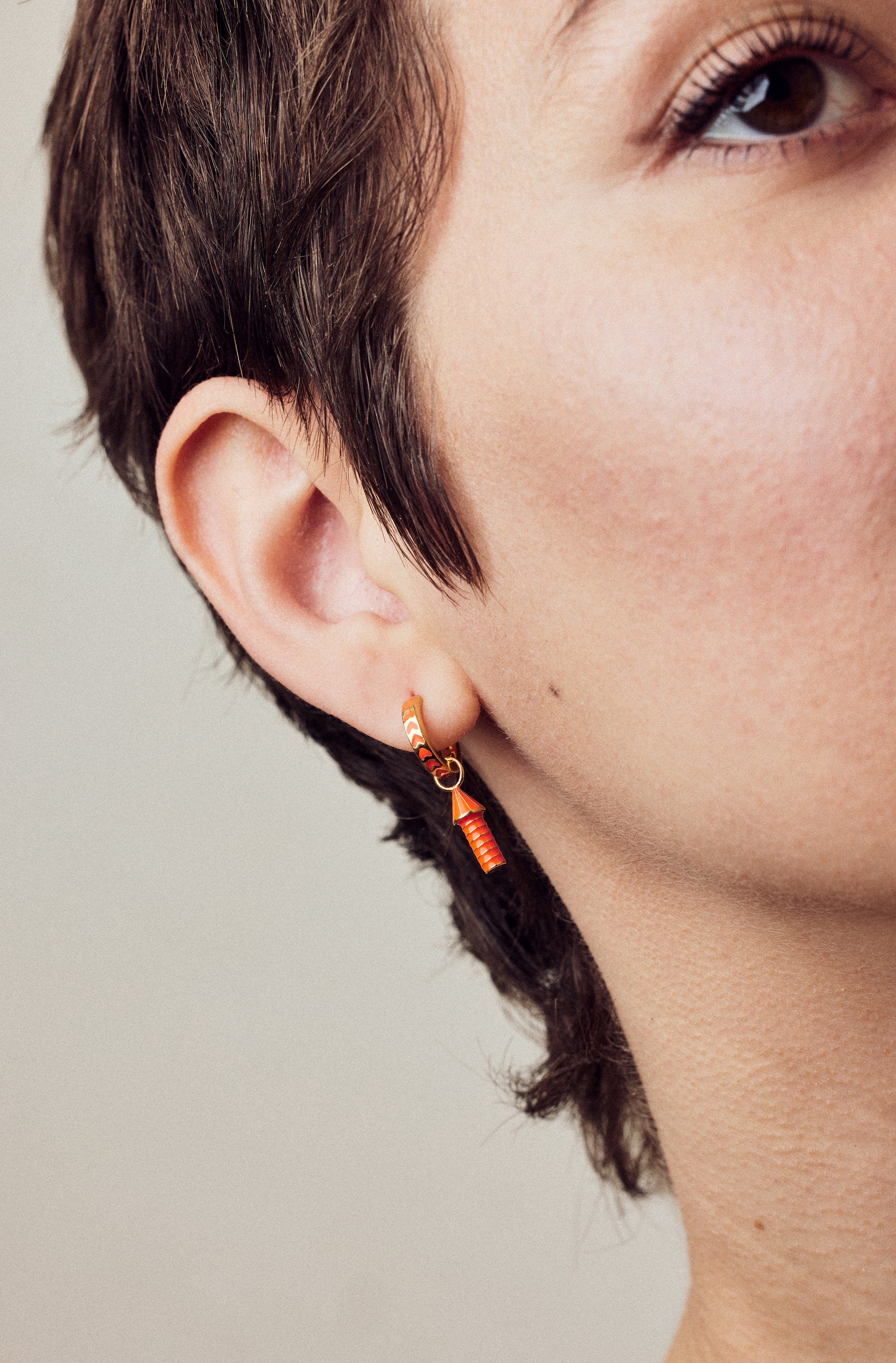 image of rocket enamel earrings in orange and gold shown close-up of ear and eye on model with white skin and short brown hair