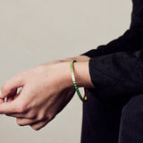 image of spark enamel bangle in green and gold on wrist of model with white skin seated with arms resting on knees