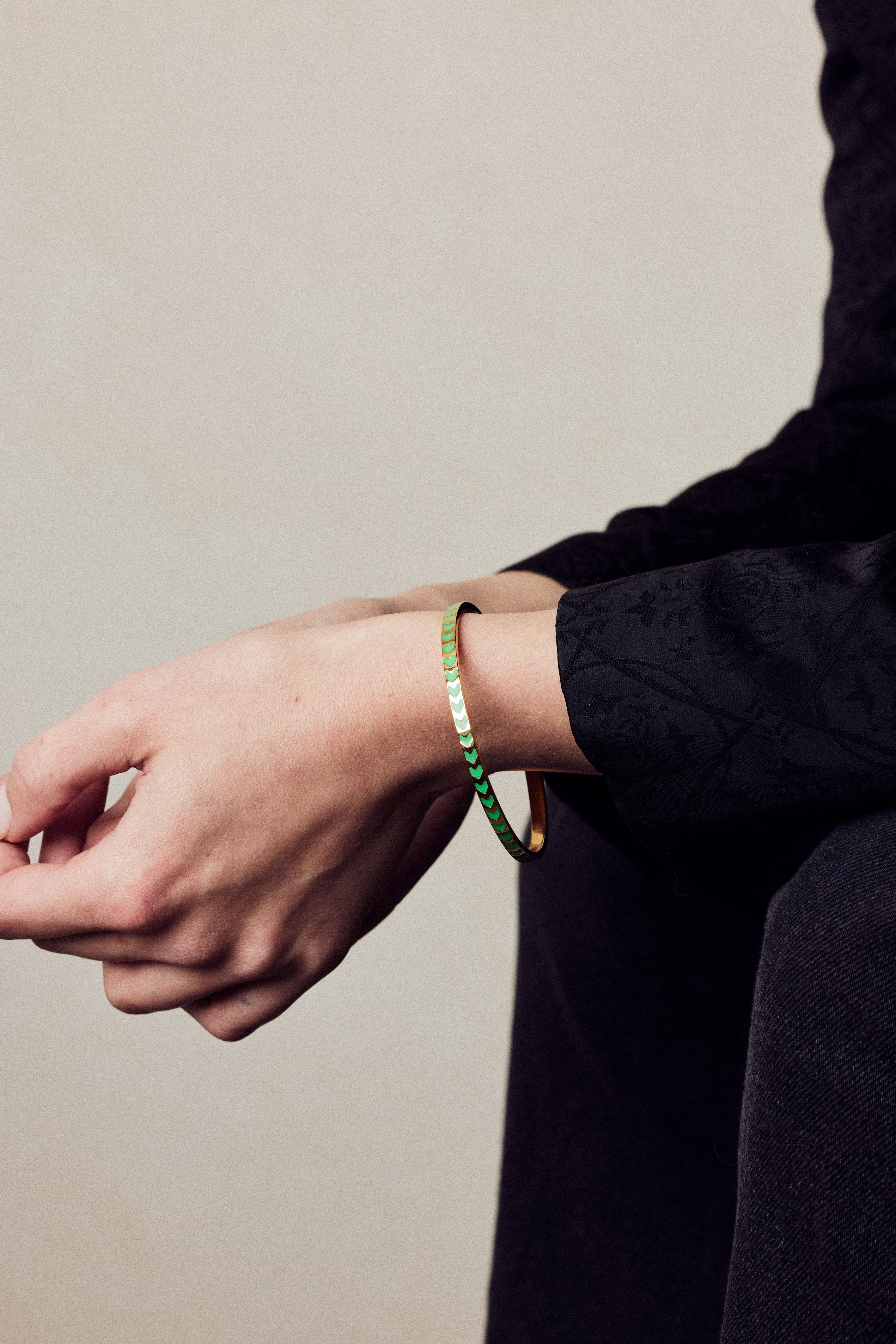 image of spark enamel bangle in green and gold on wrist of model with white skin seated with arms resting on knees