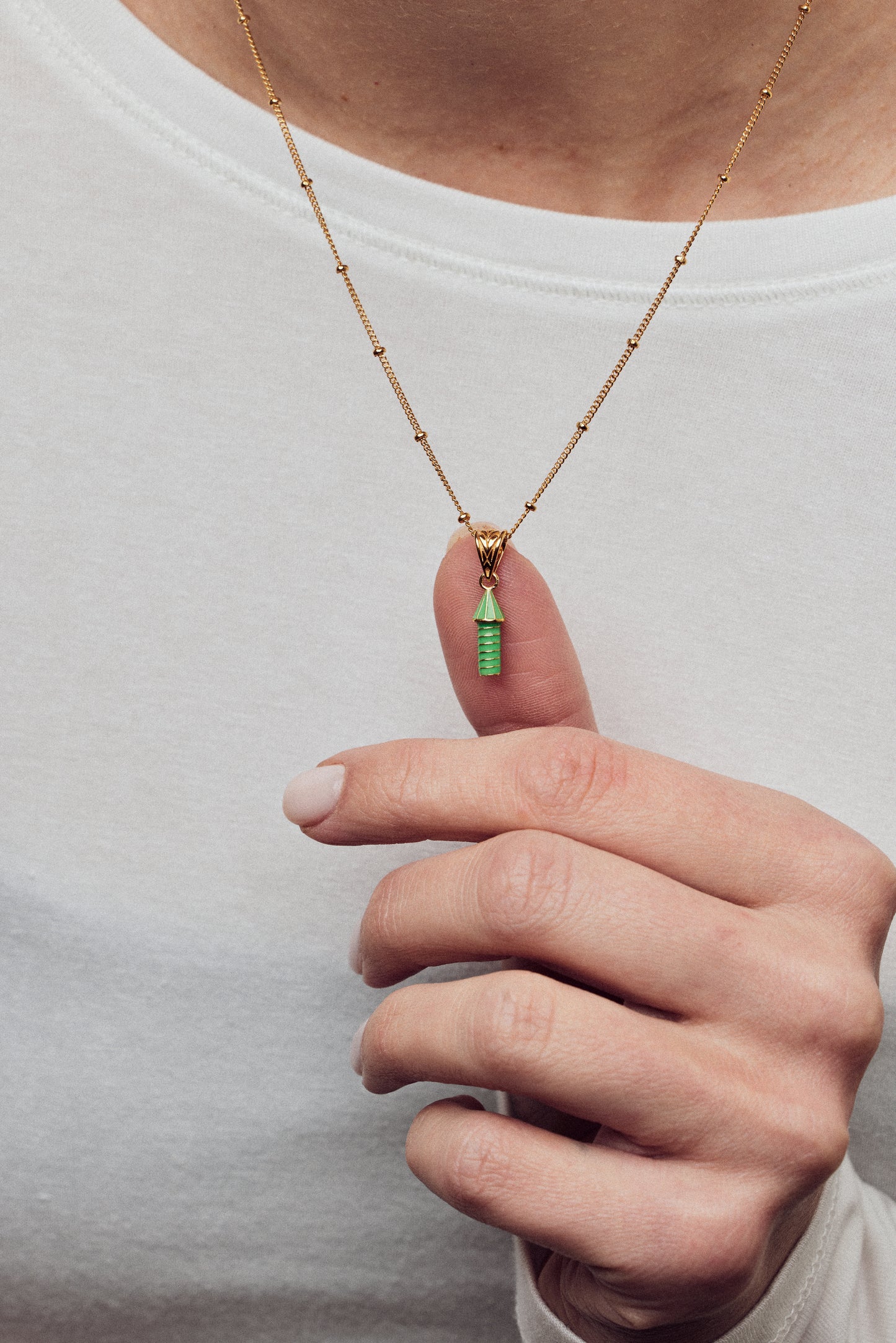 image of rocket enamel necklace in green on model in white top with thumb behind rocket charm